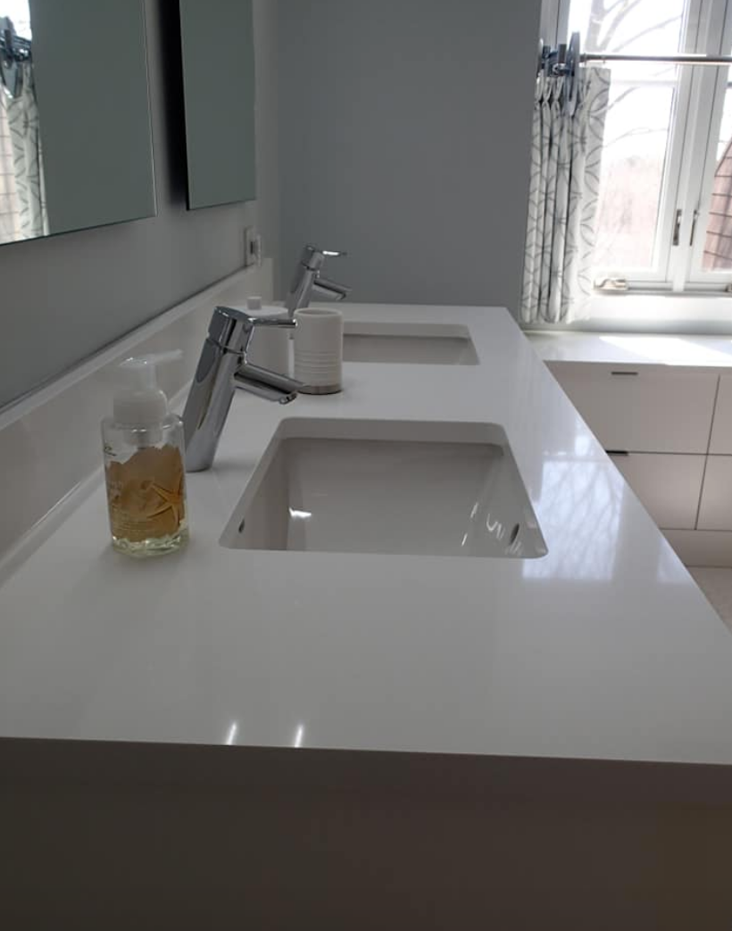 Double white sinks on a countertop in a bathroom; chrome faucets, gray walls, and window with patterned curtains.