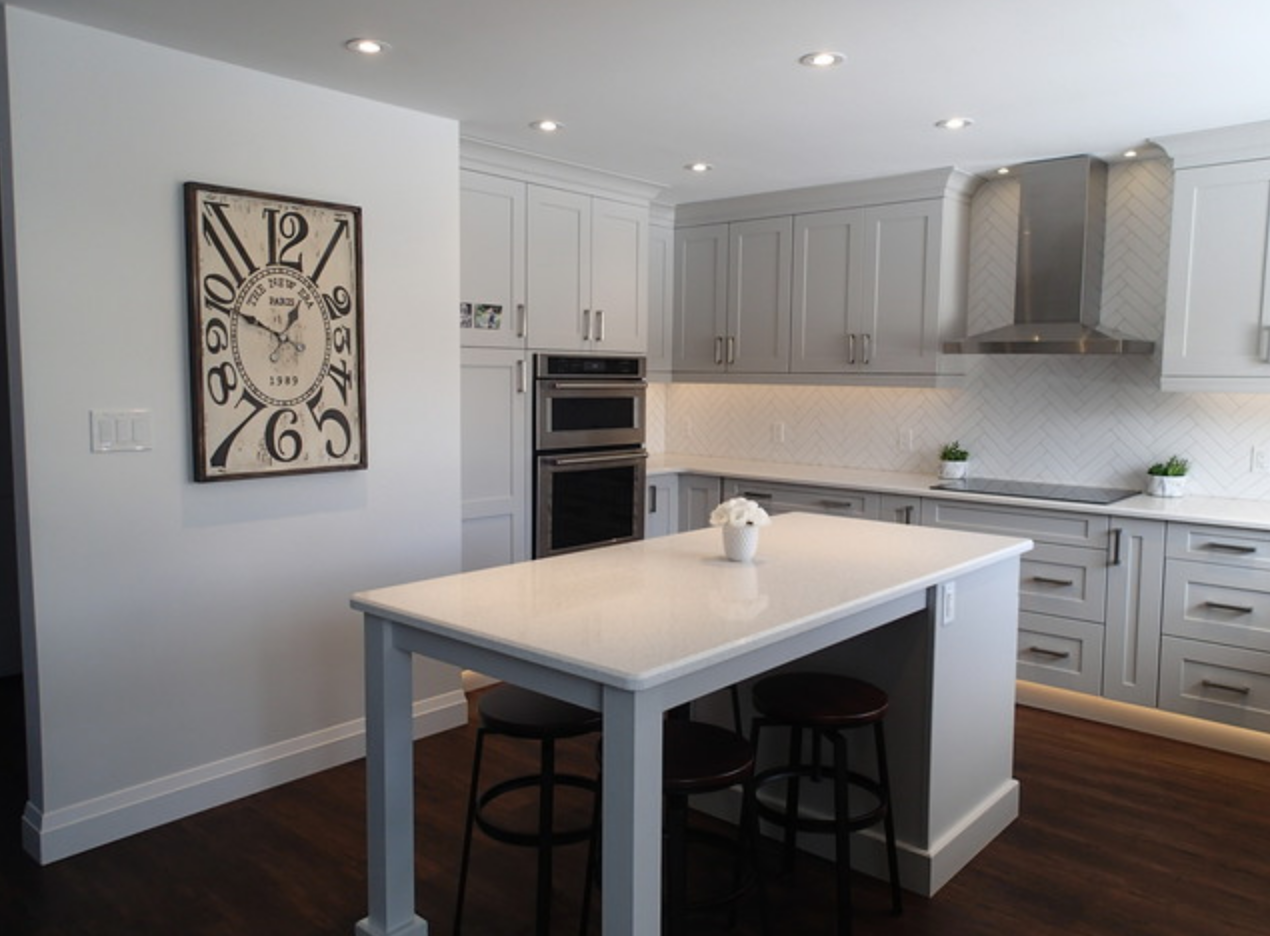 Modern kitchen with gray cabinets, white countertops, island with stools, and large clock on the wall.