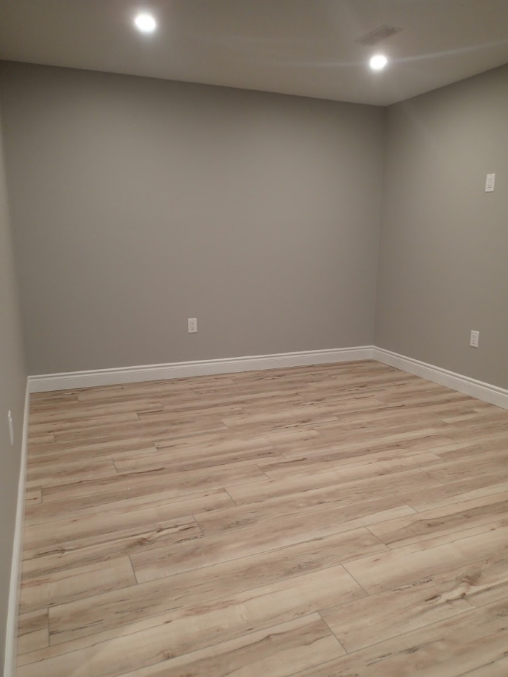 Empty room with gray walls, light wood-look flooring, and white trim. Two recessed lights illuminate the space.