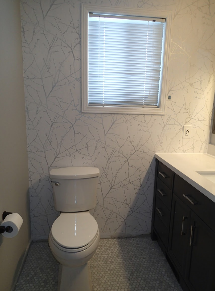 Bathroom with white toilet and sink, window with blinds, patterned wallpaper, and gray cabinets.