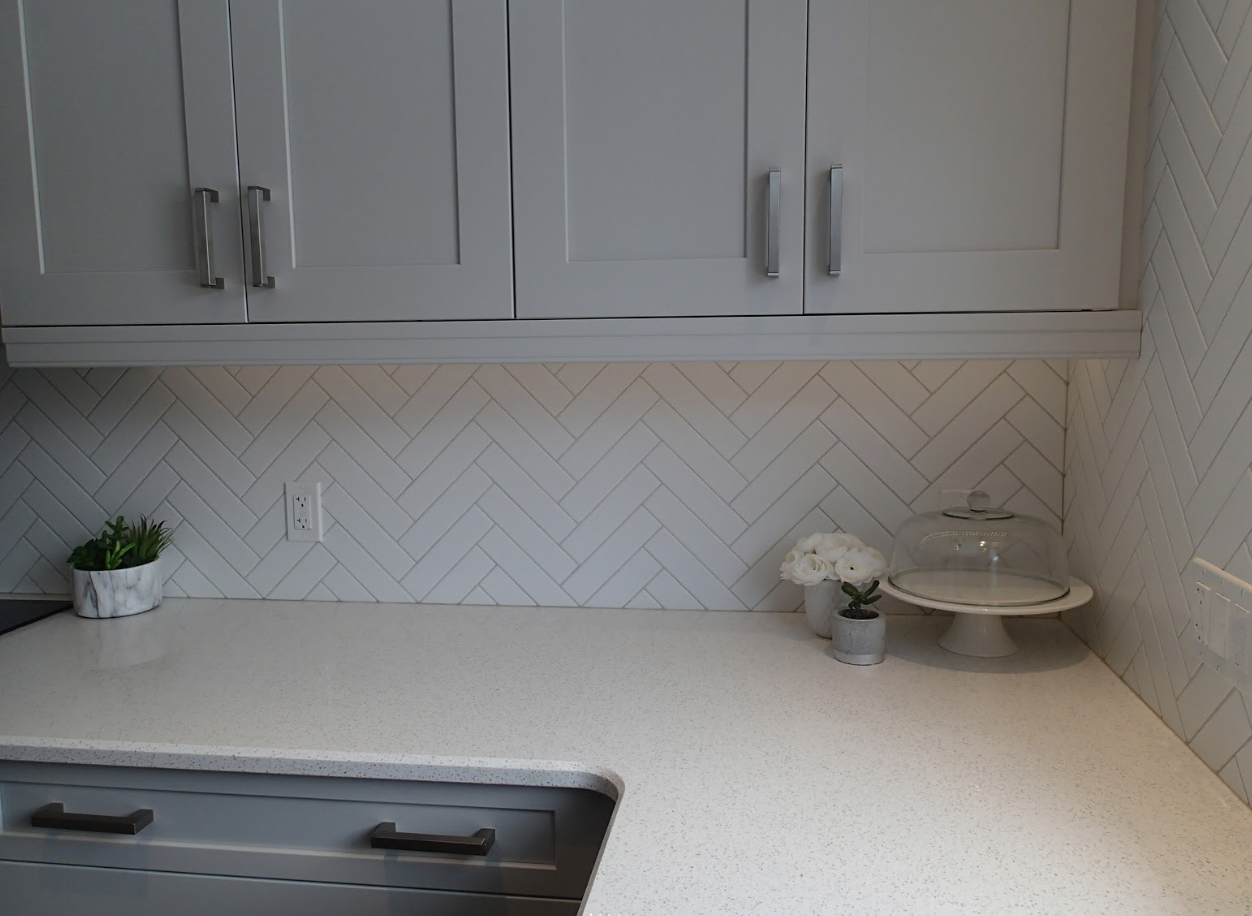 Kitchen countertop with light grey cabinets, white backsplash, and decorative items.