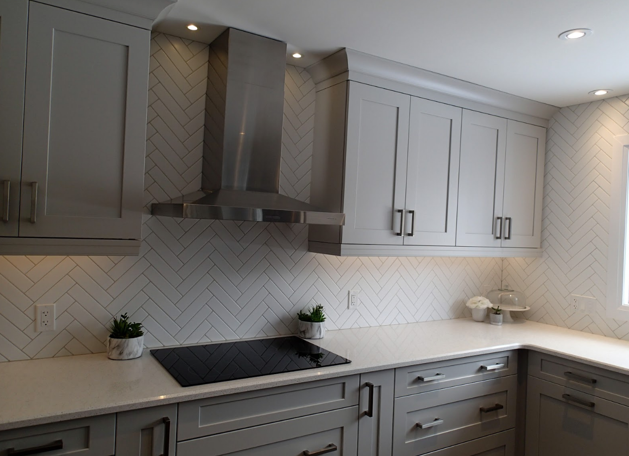 Gray kitchen with cabinets, stainless steel hood, white countertop, and herringbone tile backsplash.
