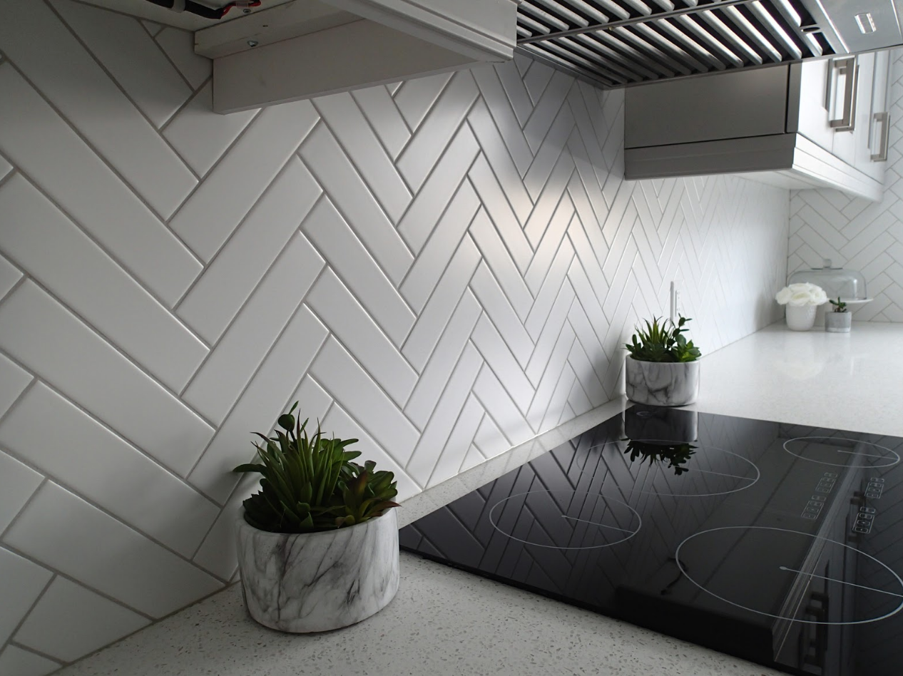 White herringbone tile backsplash in a kitchen with a black stovetop and small potted plants.