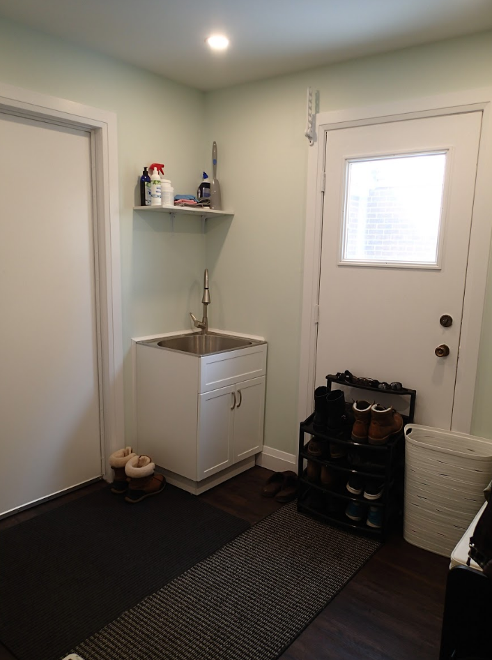 Small laundry room with sink, white door, shoes, and cleaning supplies on a shelf.