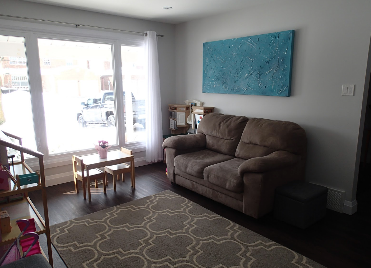 Cozy living room with dark wood floors, a gray rug, and a brown loveseat. Large window lets in light.