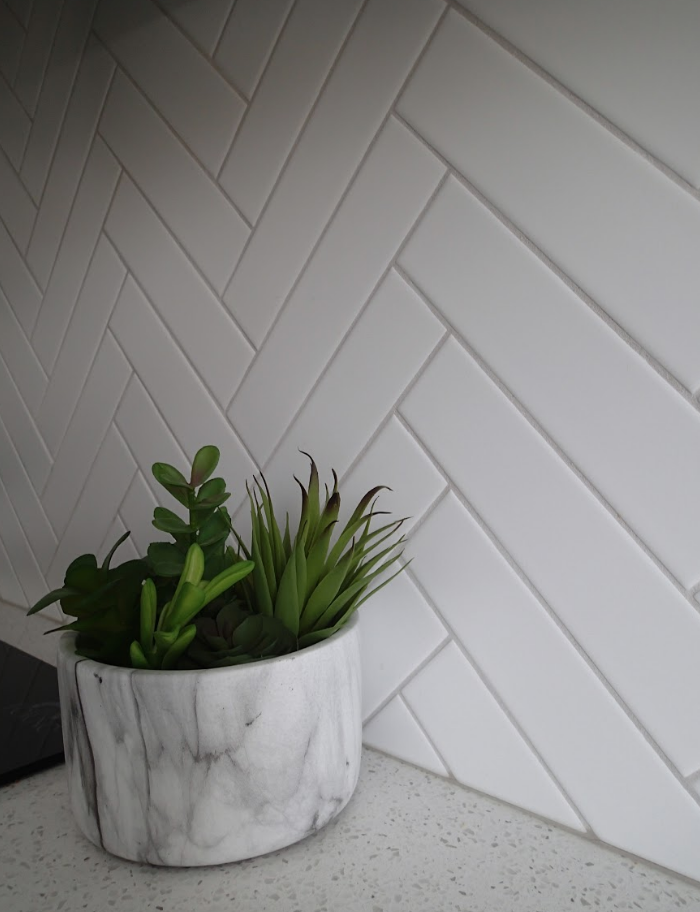 White herringbone tile backsplash with marble pot of greenery on a white countertop.