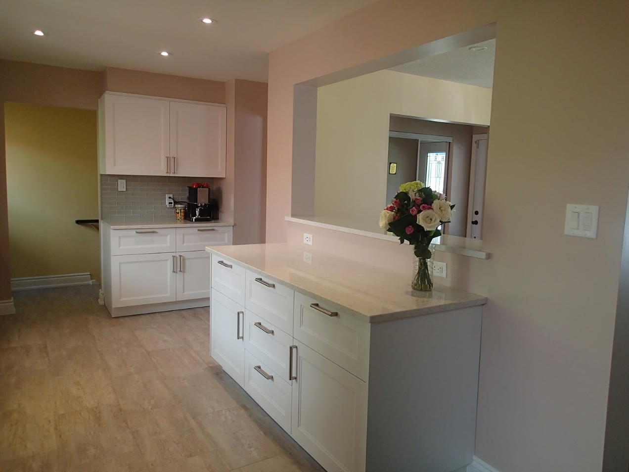 White kitchen cabinets and countertop with light-colored wood-look flooring. Pink walls and a pass-through to another room.