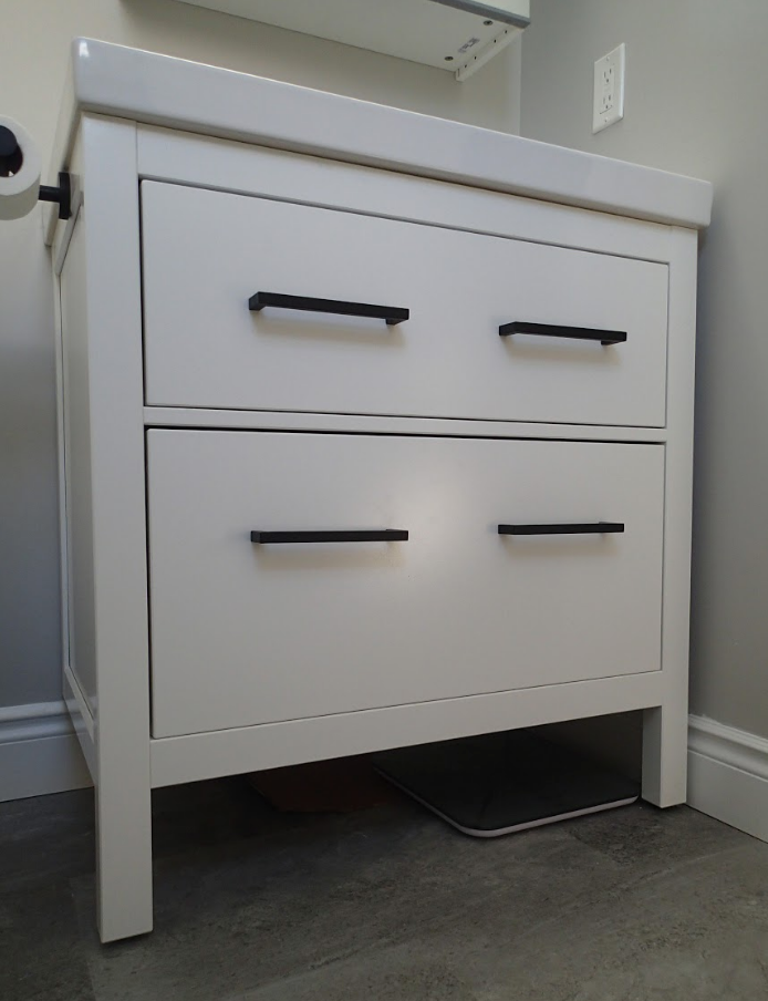 White bathroom vanity with two drawers, black handles, and a scale underneath.