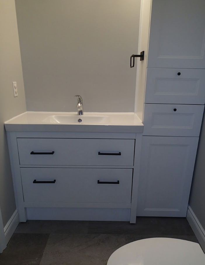 White bathroom vanity with drawers and tall storage cabinet. Gray walls and dark hardware.