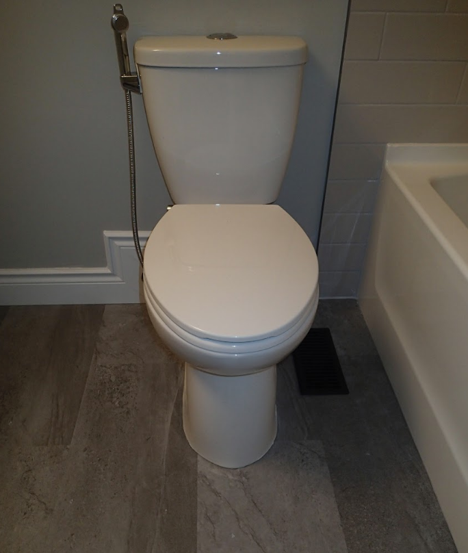 White toilet with bidet sprayer in a modern bathroom with gray flooring and walls.