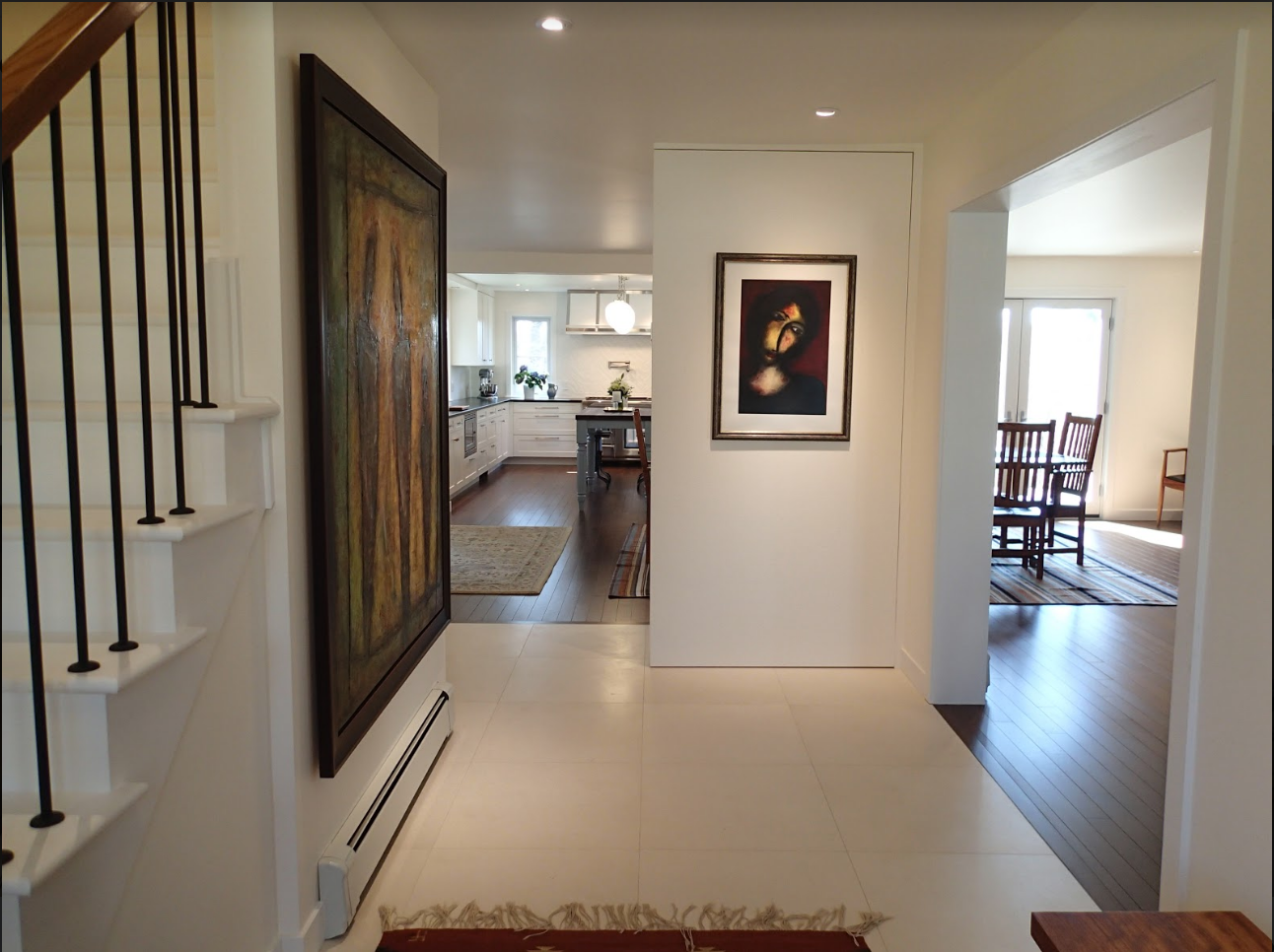 Entryway with white walls, hardwood floors, and artwork. Staircase to the left, opening to a kitchen and dining room.