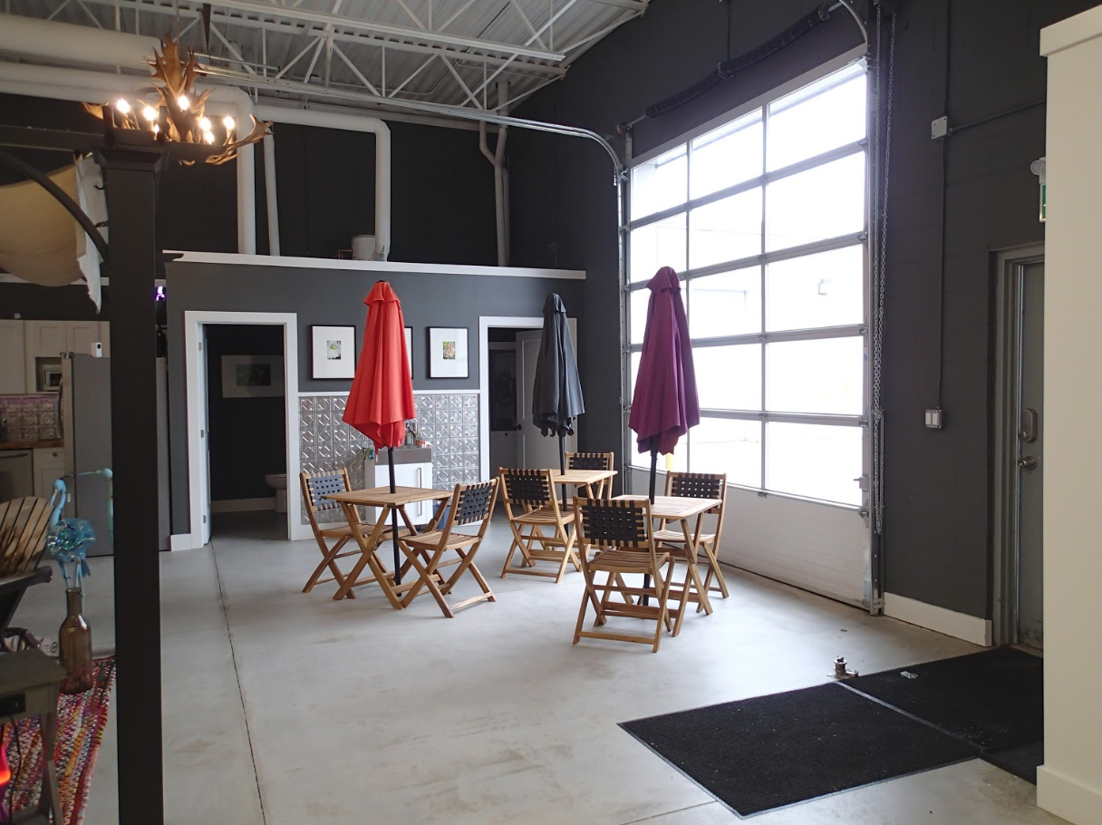 Indoor seating area with tables, chairs, umbrellas, and a large window. Dark walls, and a light-colored floor.