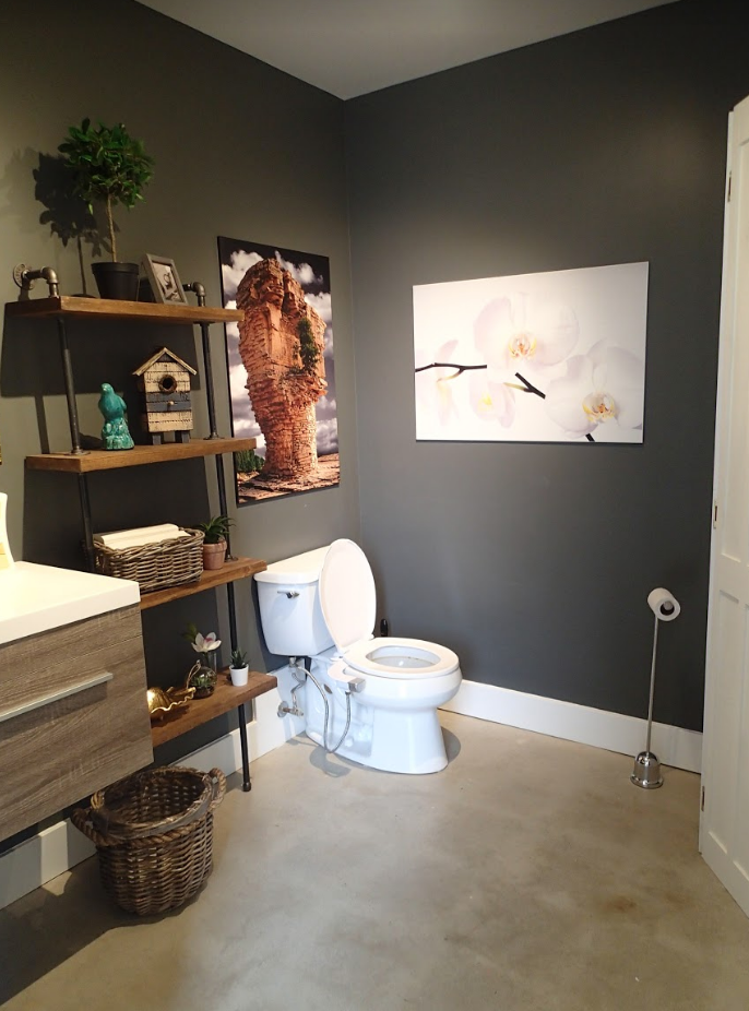 A small bathroom with gray walls, toilet, and artwork. A wooden shelf displays decor; a white cabinet is on the right.