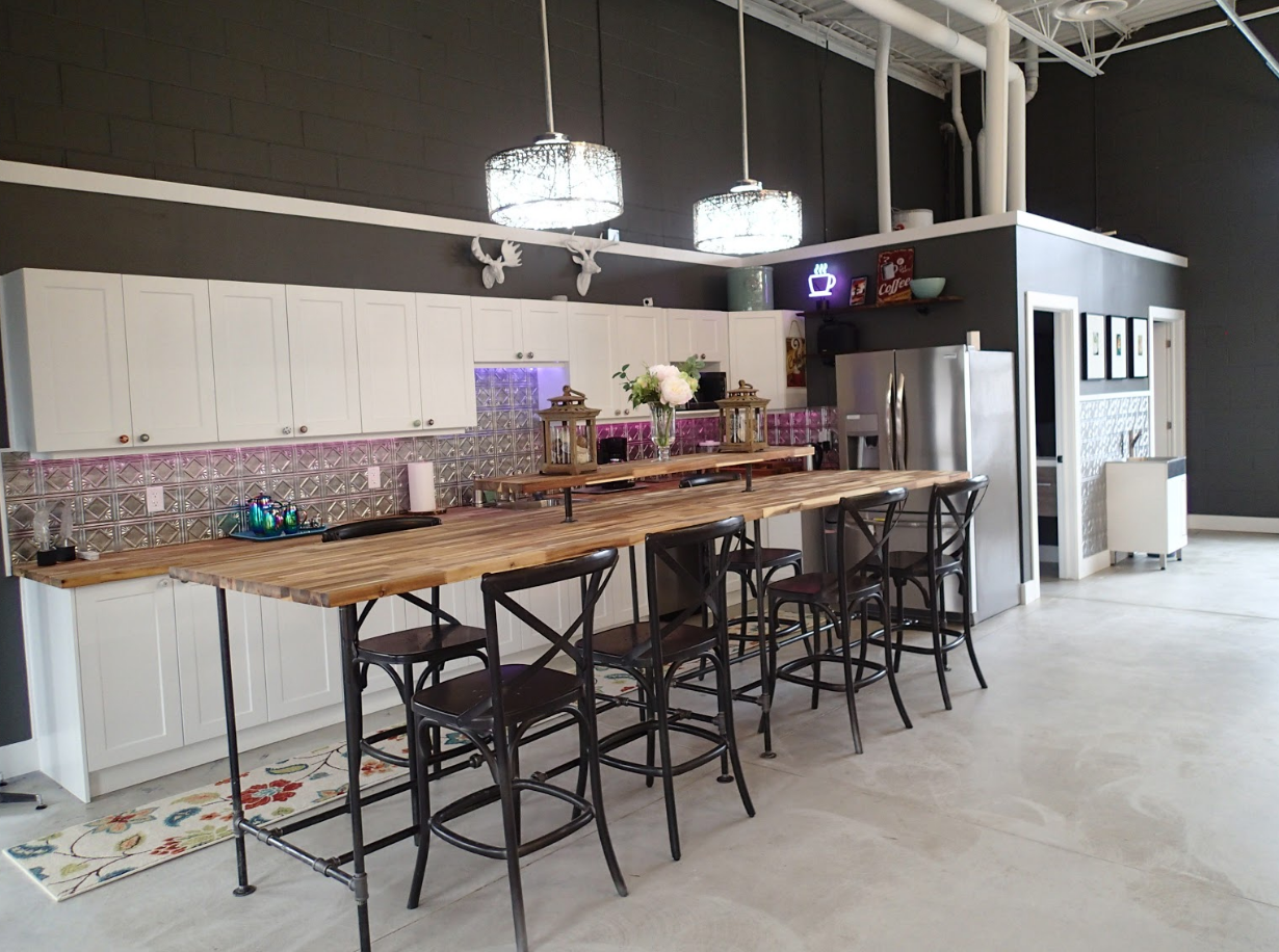 Modern kitchen with white cabinets, long wood island with metal chairs, stainless steel refrigerator, and dark gray walls.