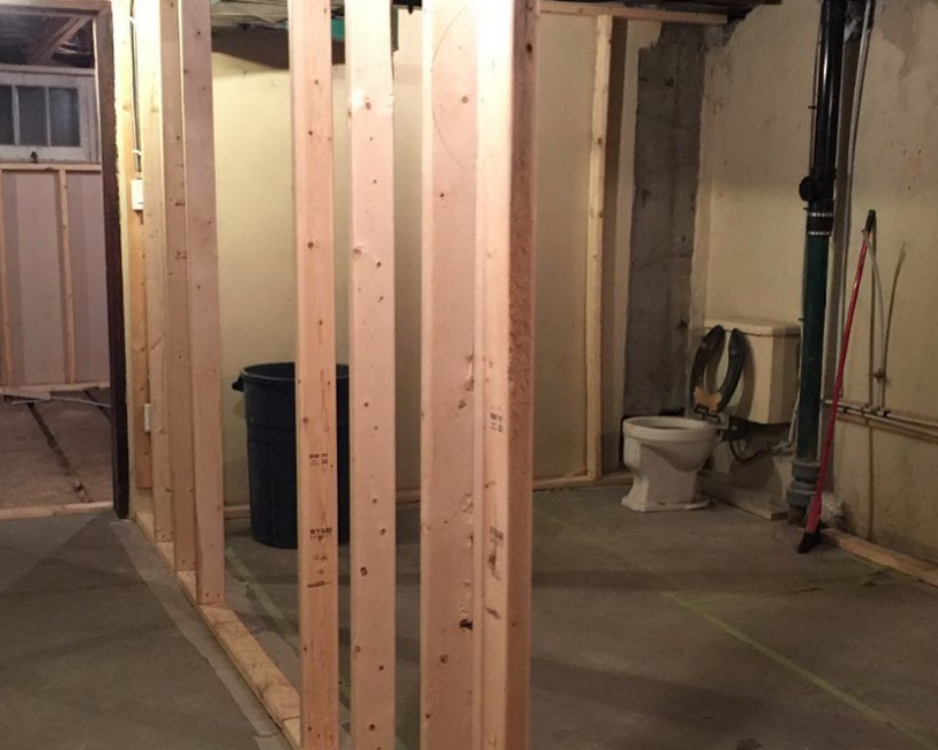 Basement renovation: Wooden studs framing walls. Toilet, trash can, and unfinished concrete floor visible.