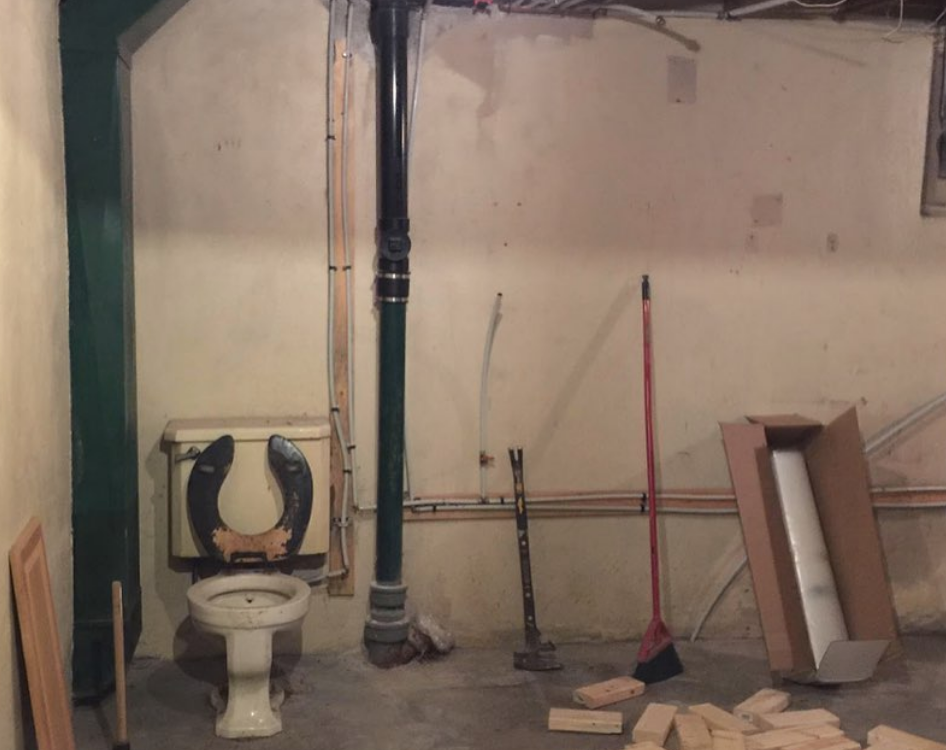 Old toilet in unfinished basement; black pipe, exposed framing, some tools, and a broom.