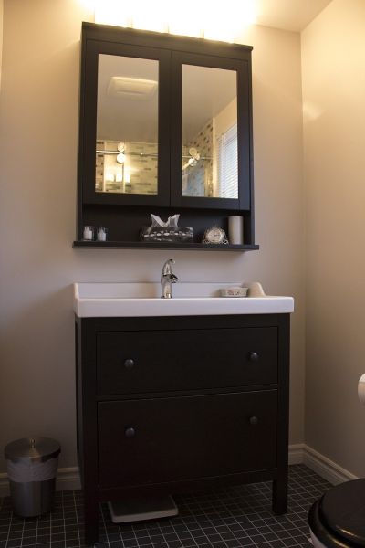 Dark vanity with mirror cabinet, shelf, white sink, and trash can in a bathroom with dark tiled floor and beige walls.