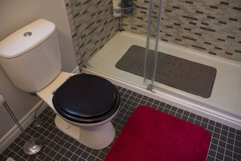 Toilet with black seat, red bath mat, and shower with dark gray mat in a tiled bathroom.