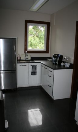 Small, white kitchen with stainless steel fridge, dark countertops, and a window.