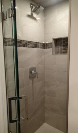 Shower with glass door, neutral-toned tile, dark tile border, built-in shelf, and chrome fixtures.