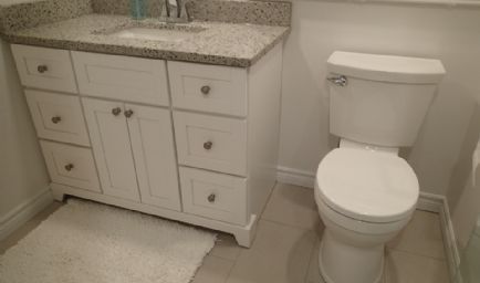 White bathroom with vanity, granite countertop, toilet, and white rug.