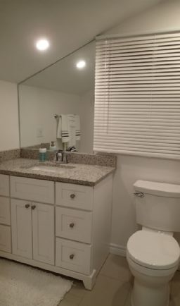 Bathroom with white vanity, toilet, and window with blinds. Mirror and countertop with granite finish.