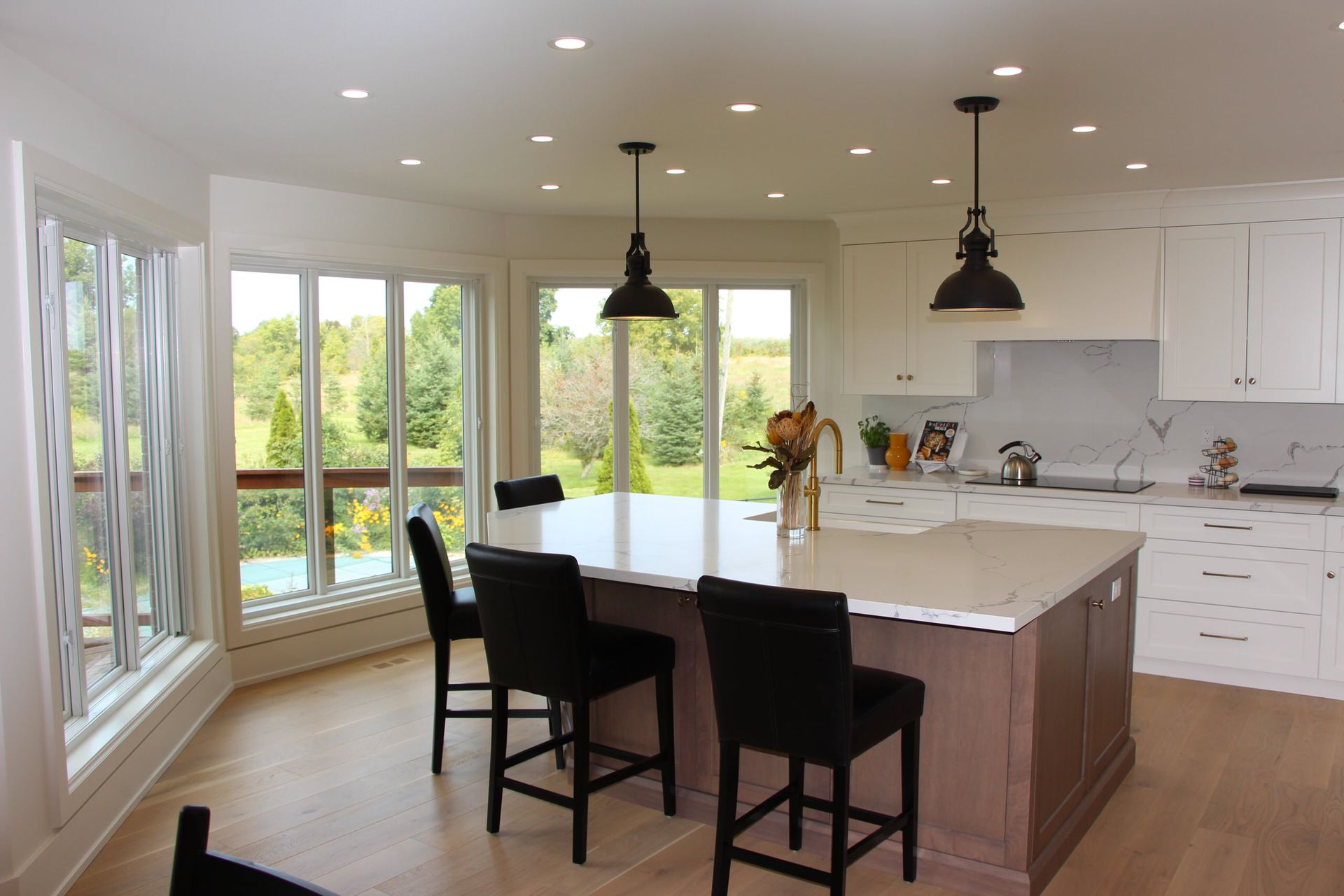 Bright kitchen with island, black bar stools, and large windows overlooking a green landscape.