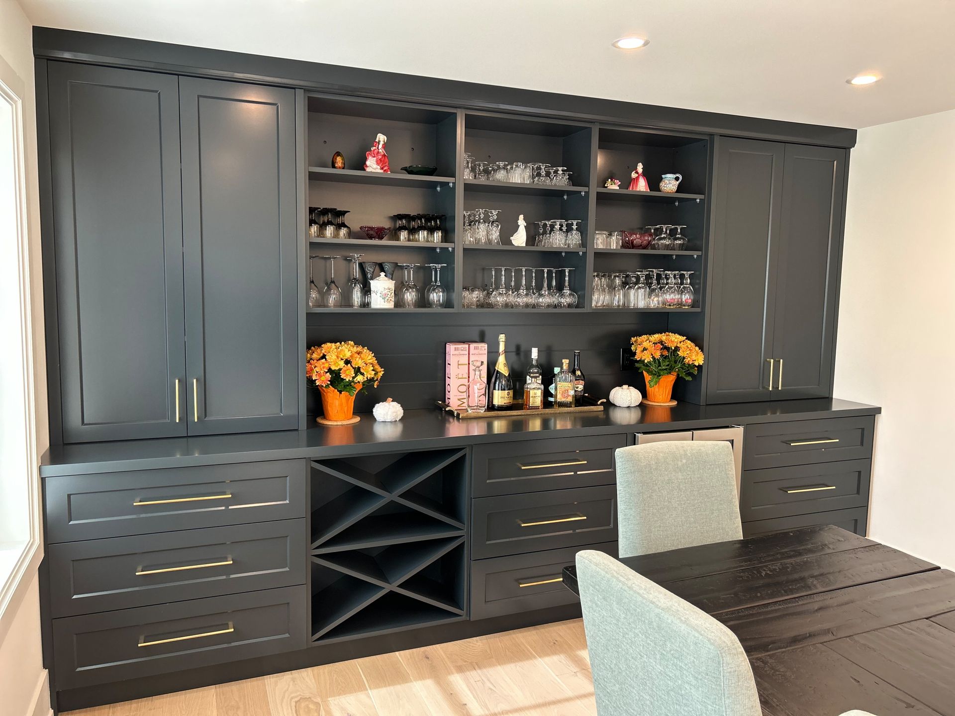 Dark gray bar cabinet with gold hardware, wine rack, and glassware; set next to a dining table.