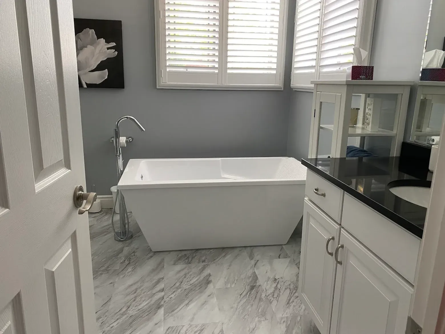 Bathroom with white freestanding tub, grey walls, shutters, white cabinets, and marble flooring.