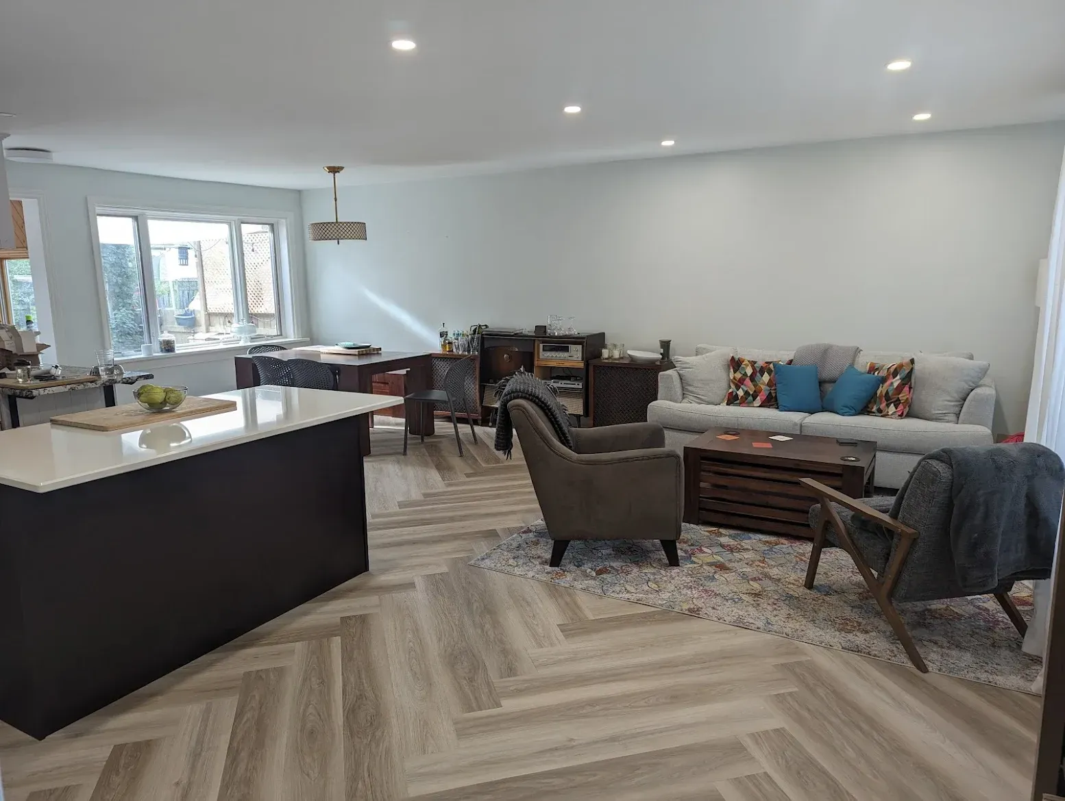 Open-plan living space with gray couch, chairs, kitchen island, and herringbone floors.