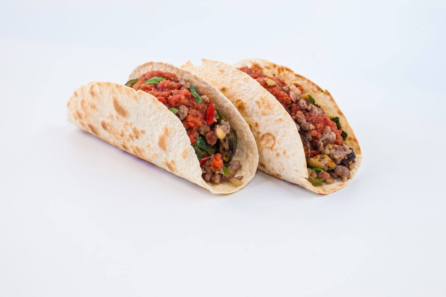 Tres tacos rellenos de carne y verduras sobre una superficie blanca.