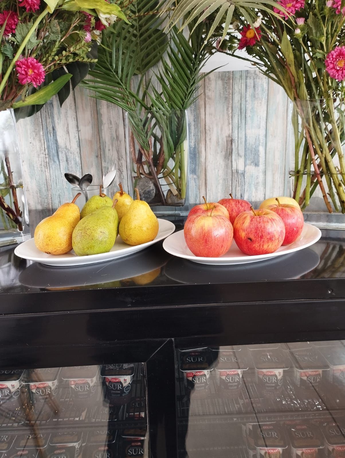 Peras y manzanas en platos blancos sobre una superficie negra. Flores y plantas al fondo.