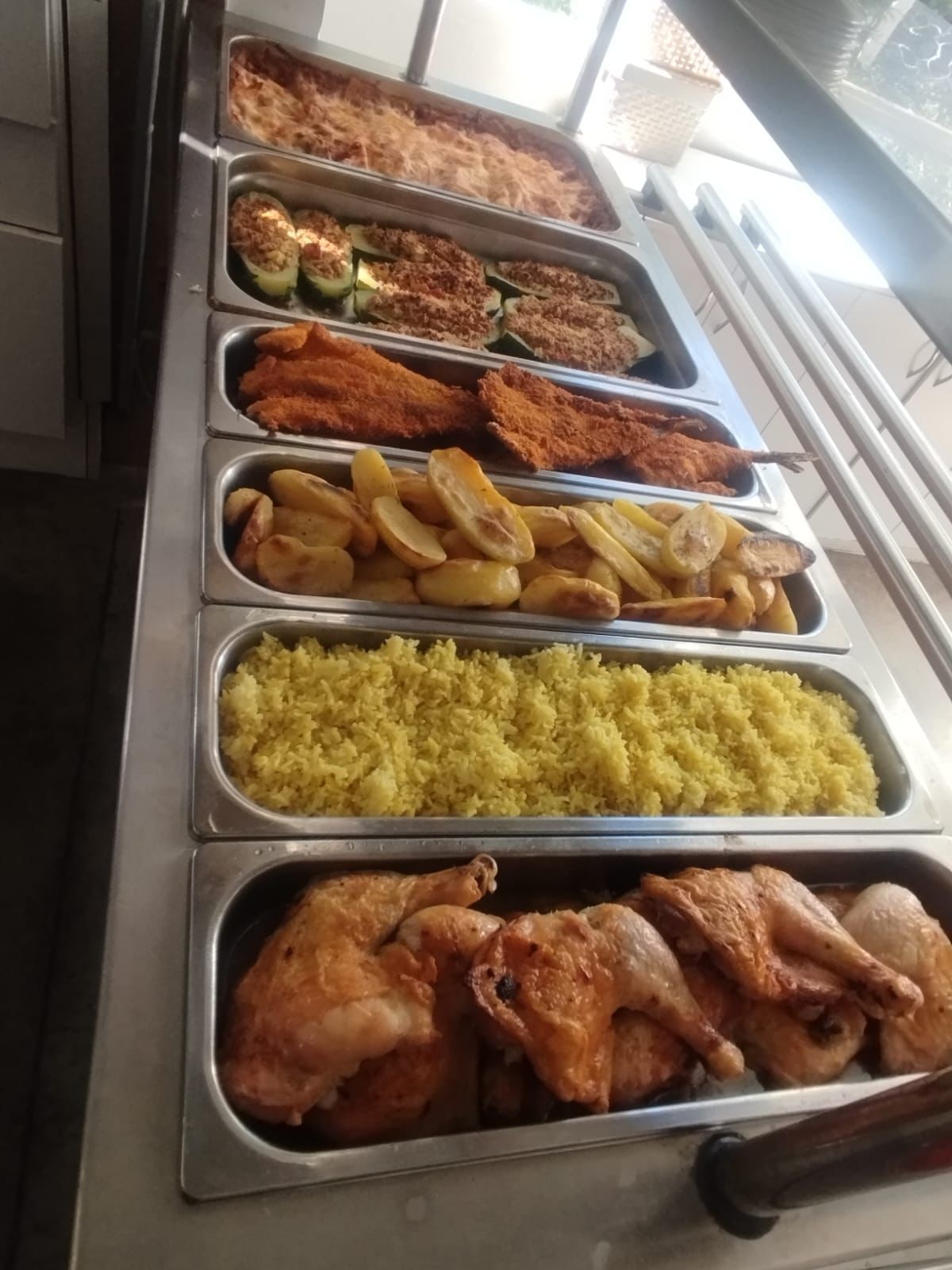 Exhibición de alimentos cocinados en forma de buffet: pollo, arroz, patatas, chuletas, calabacín y guisos.