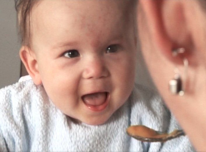 Ein lachendes Baby schaut eine erwachsene Person an, die es mit einem Löffel Brei füttert.
