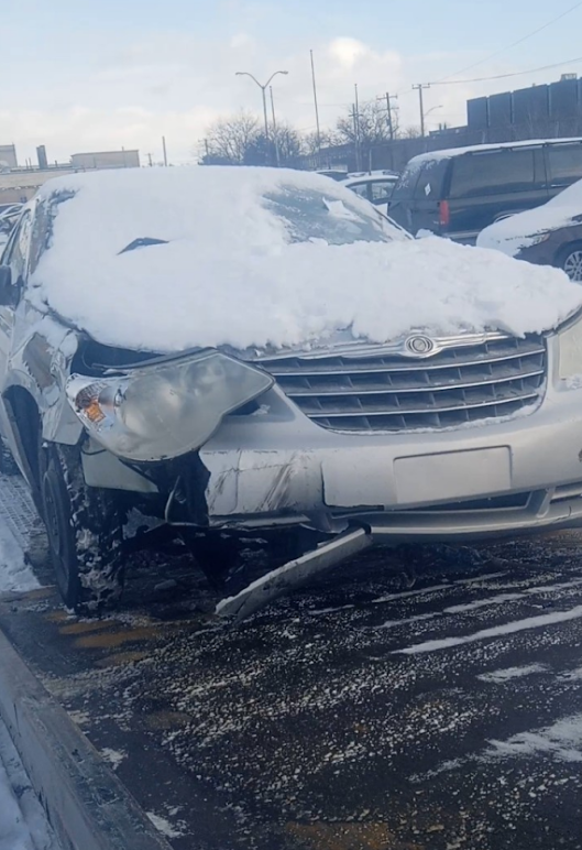 Damaged silver Chrysler covered in snow, parked outside on a winter day.
