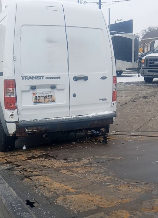 White Transit van with damaged rear bumper parked outdoors on a worn, brown surface; snow present.