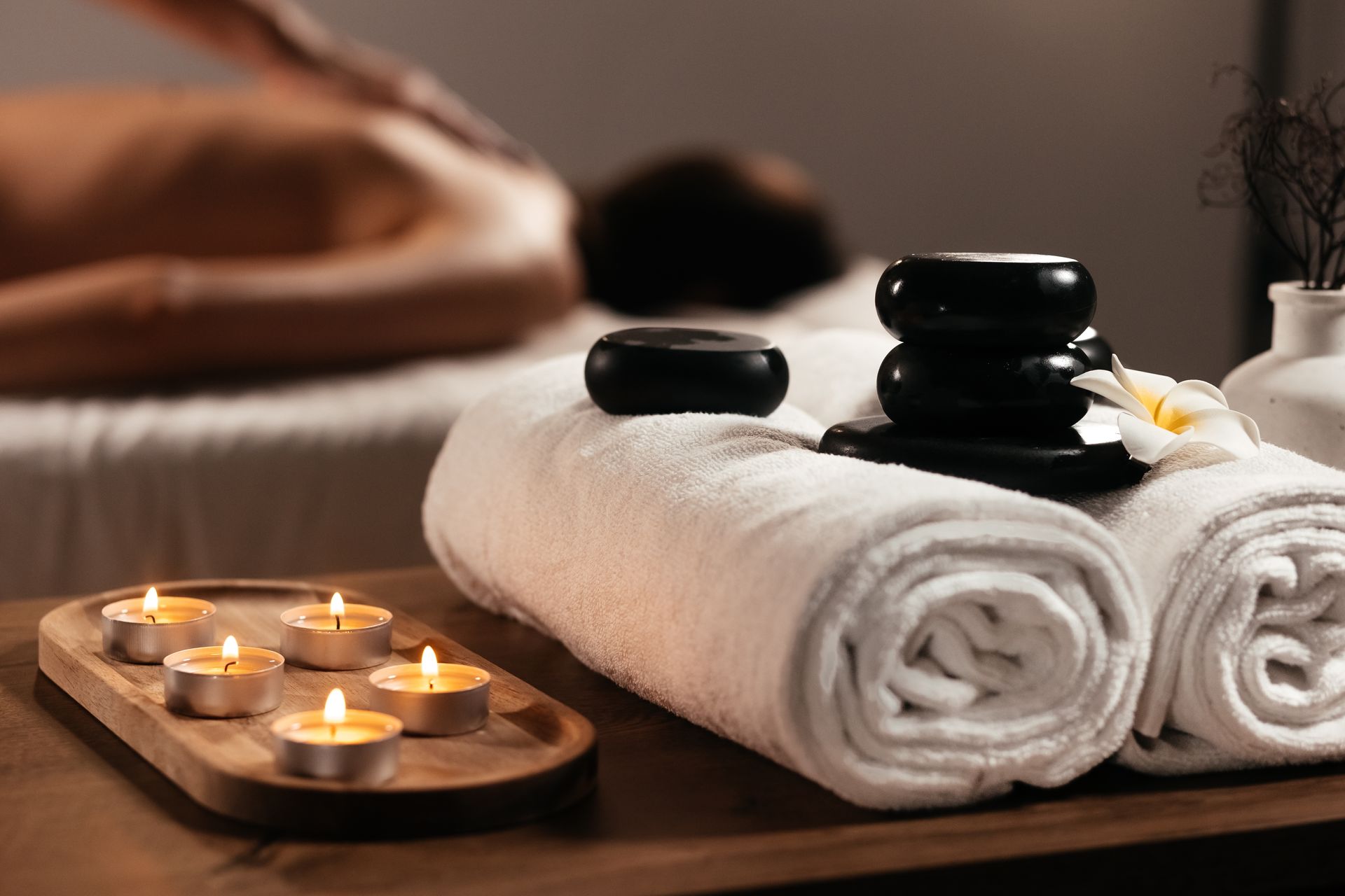 Spa scene with rolled white towels, lit candles, and hot stones, with a person receiving a massage in the background.