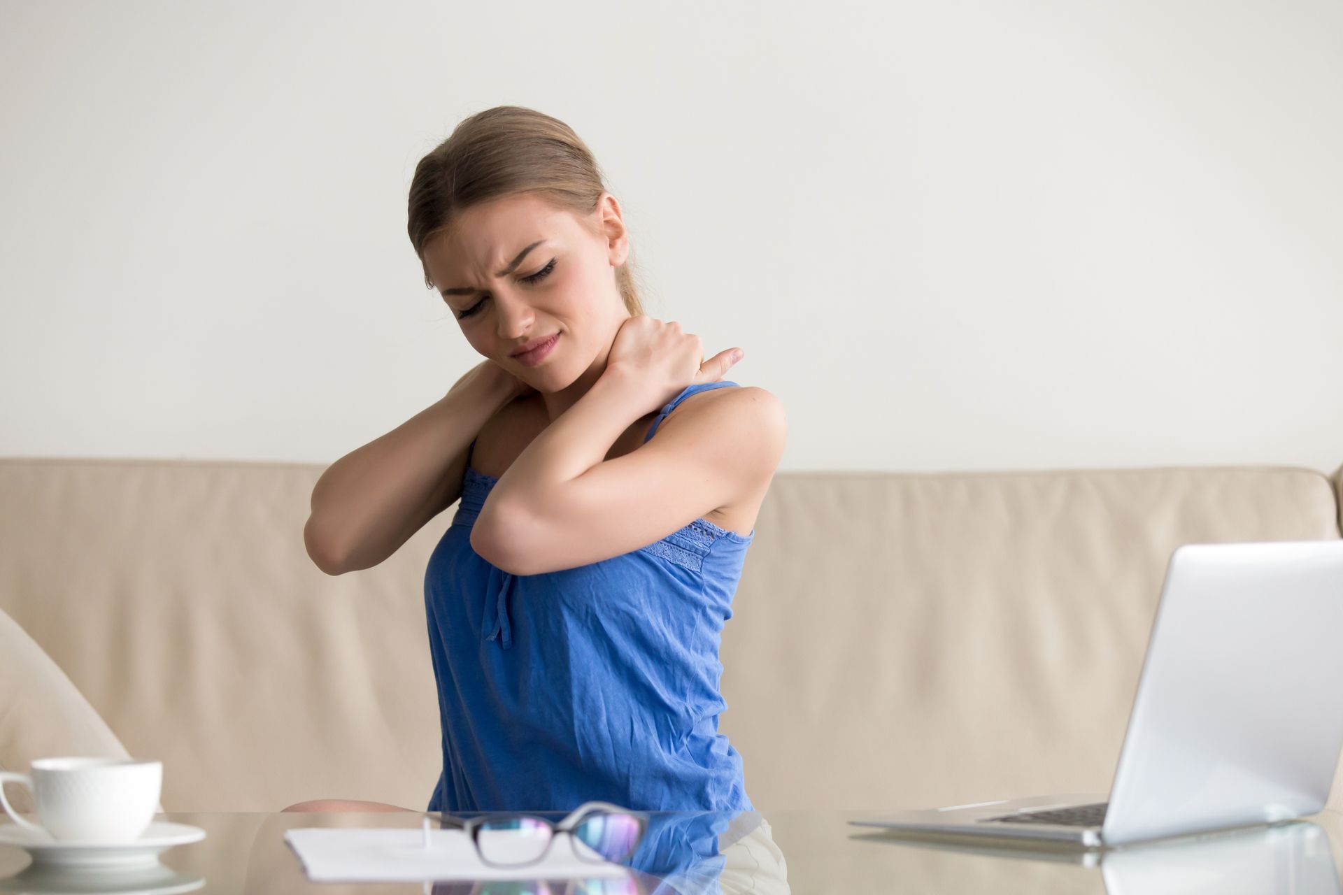 Woman in blue top grimaces, clutching her neck in pain, seated at a table with a laptop and coffee.