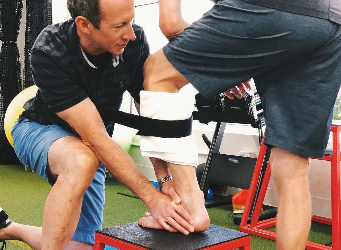 A man is kneeling down to help another man stretch his leg