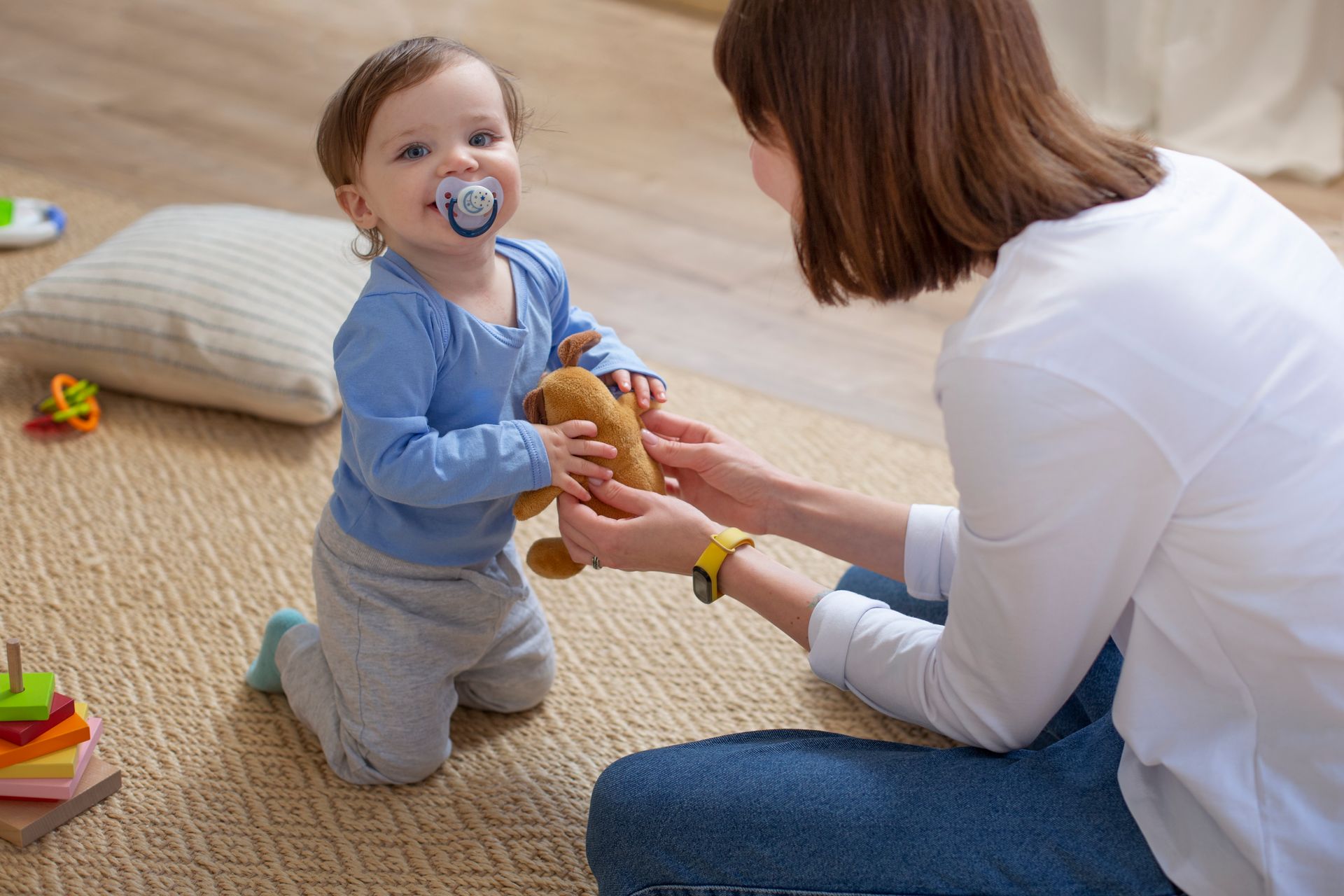 A woman is kneeling down next to a baby with a pacifier in his mouth.