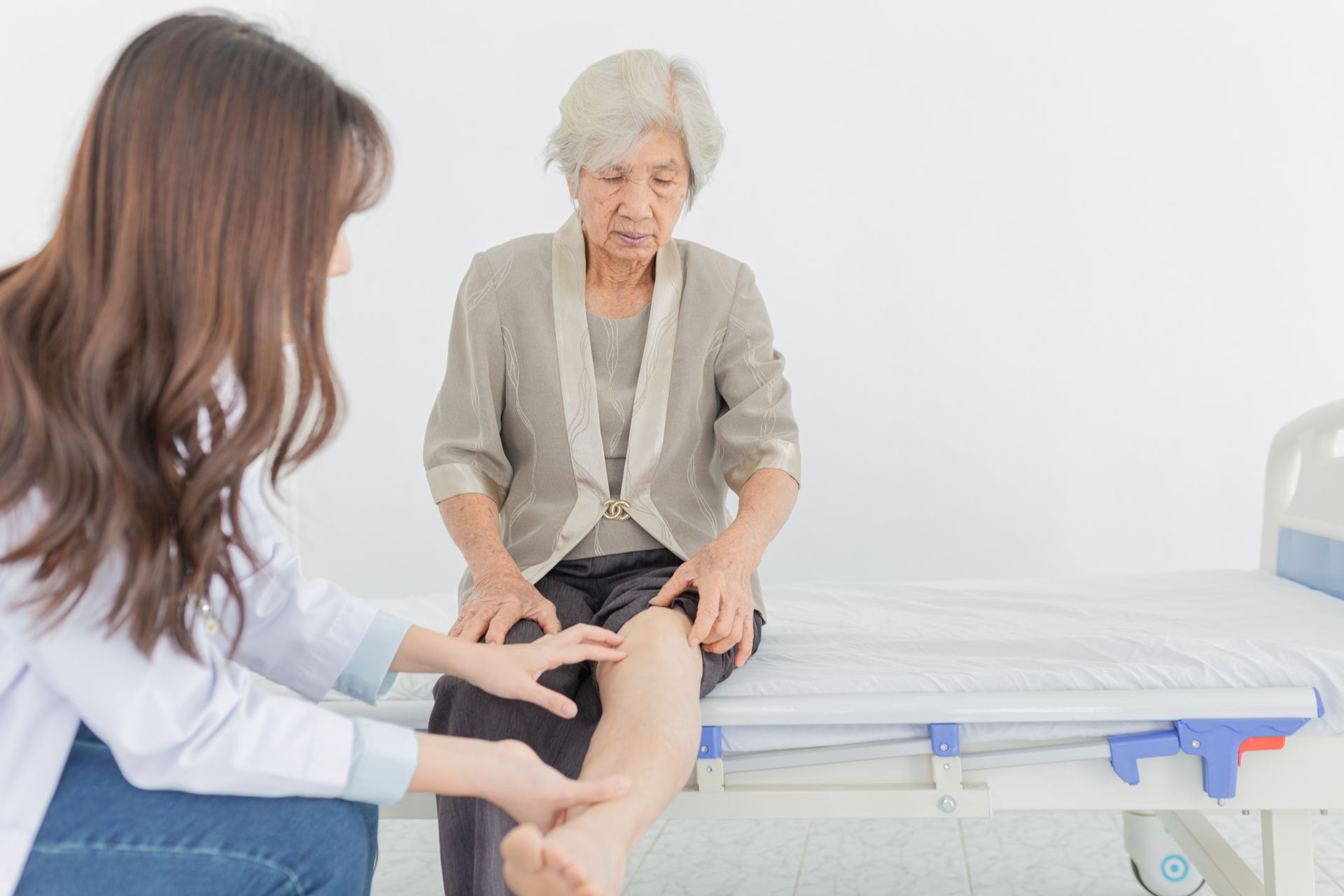 An elderly woman is sitting on a hospital bed while a doctor examines her leg.