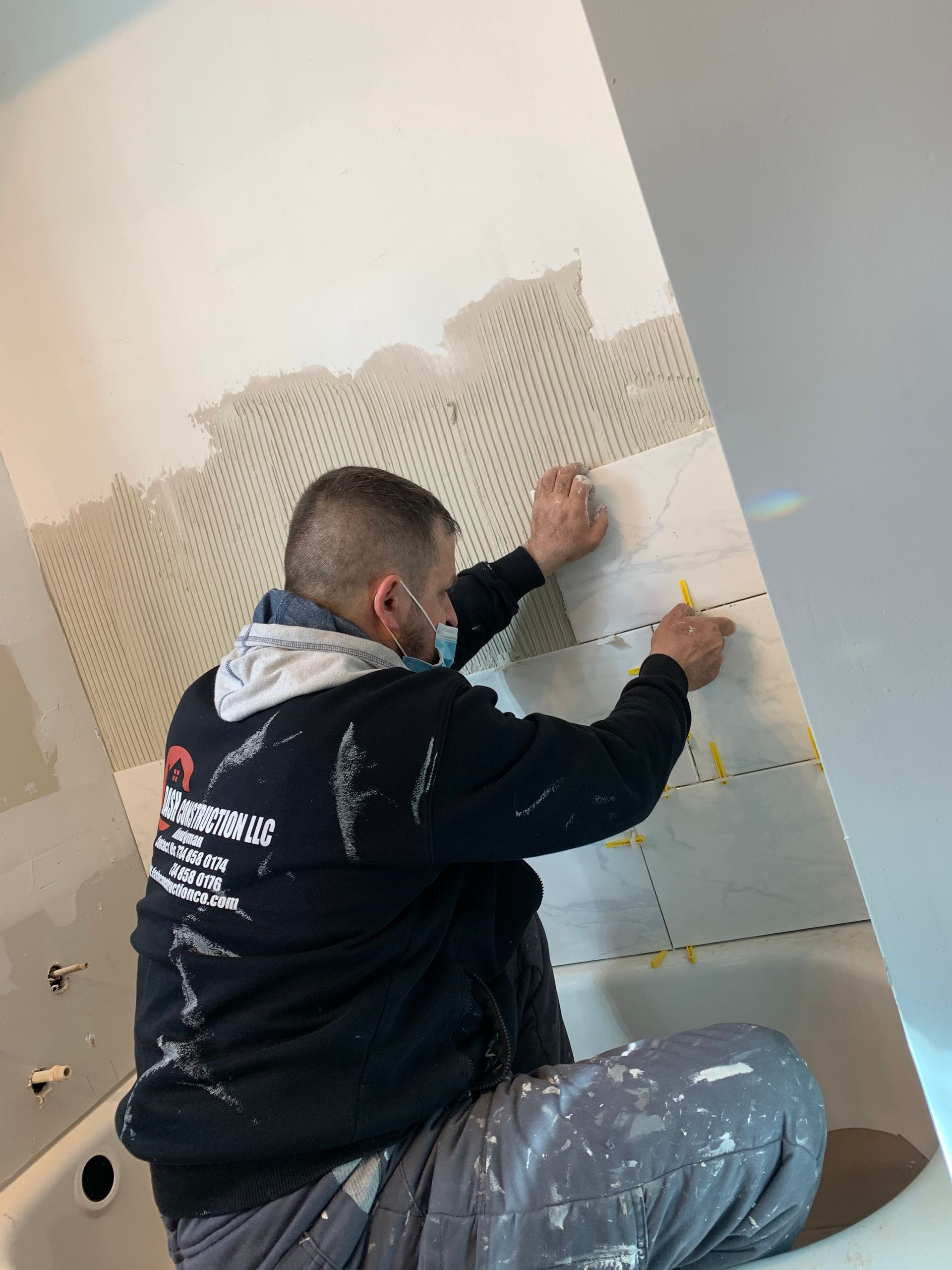 A person tiling a bathroom wall, using yellow spacers. The worker is wearing a mask and dark hoodie, in front of a white wall.