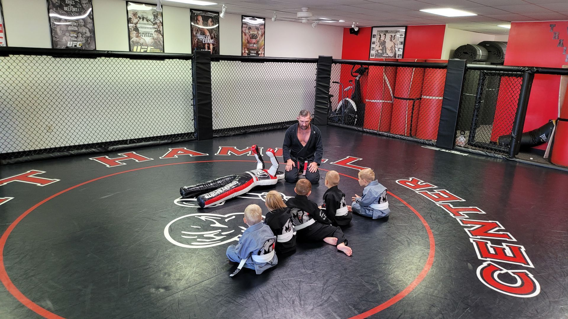 A group of children are sitting on a wrestling mat in a gym.