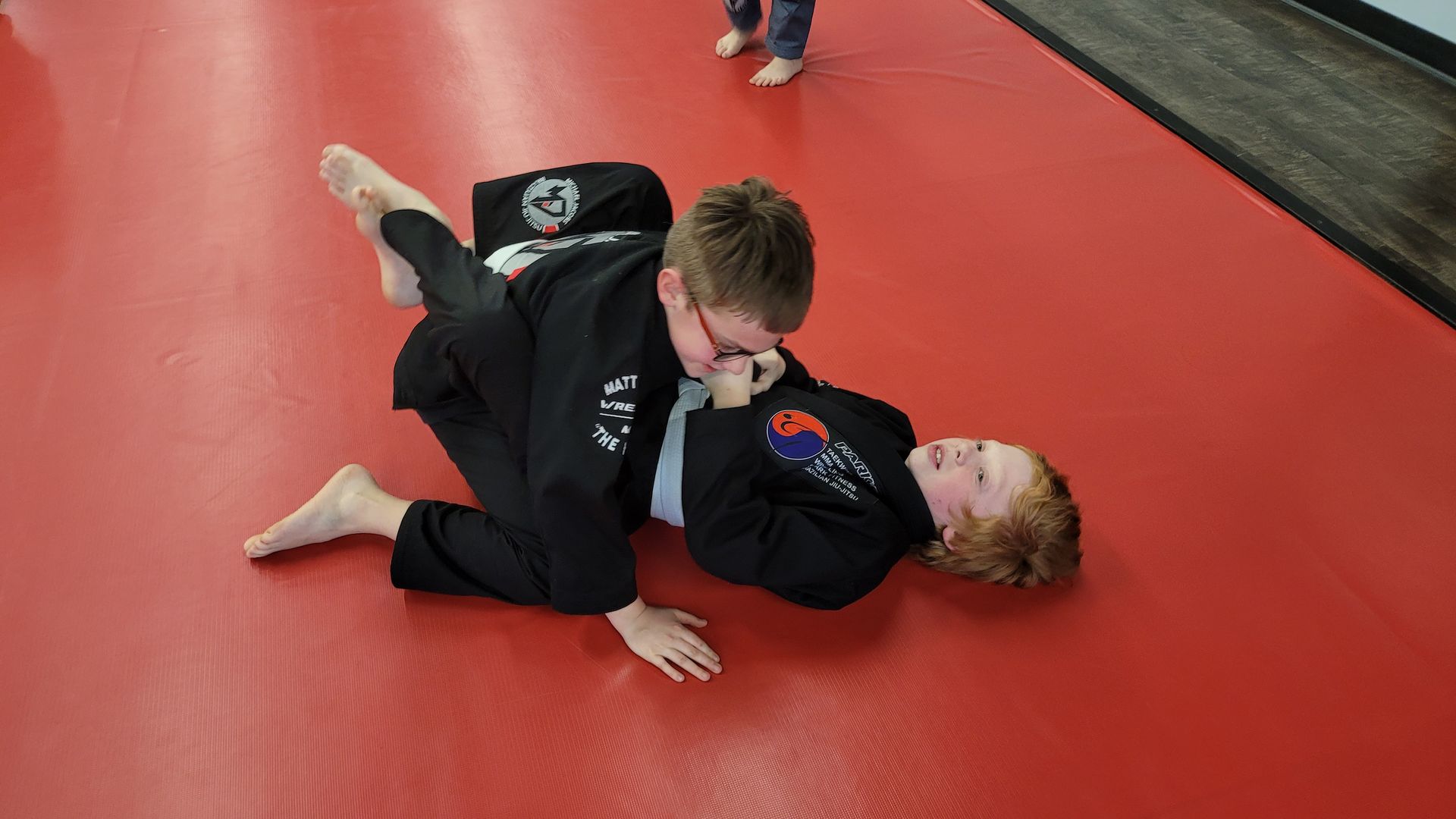 Two young boys are practicing jiu jitsu on a red mat.