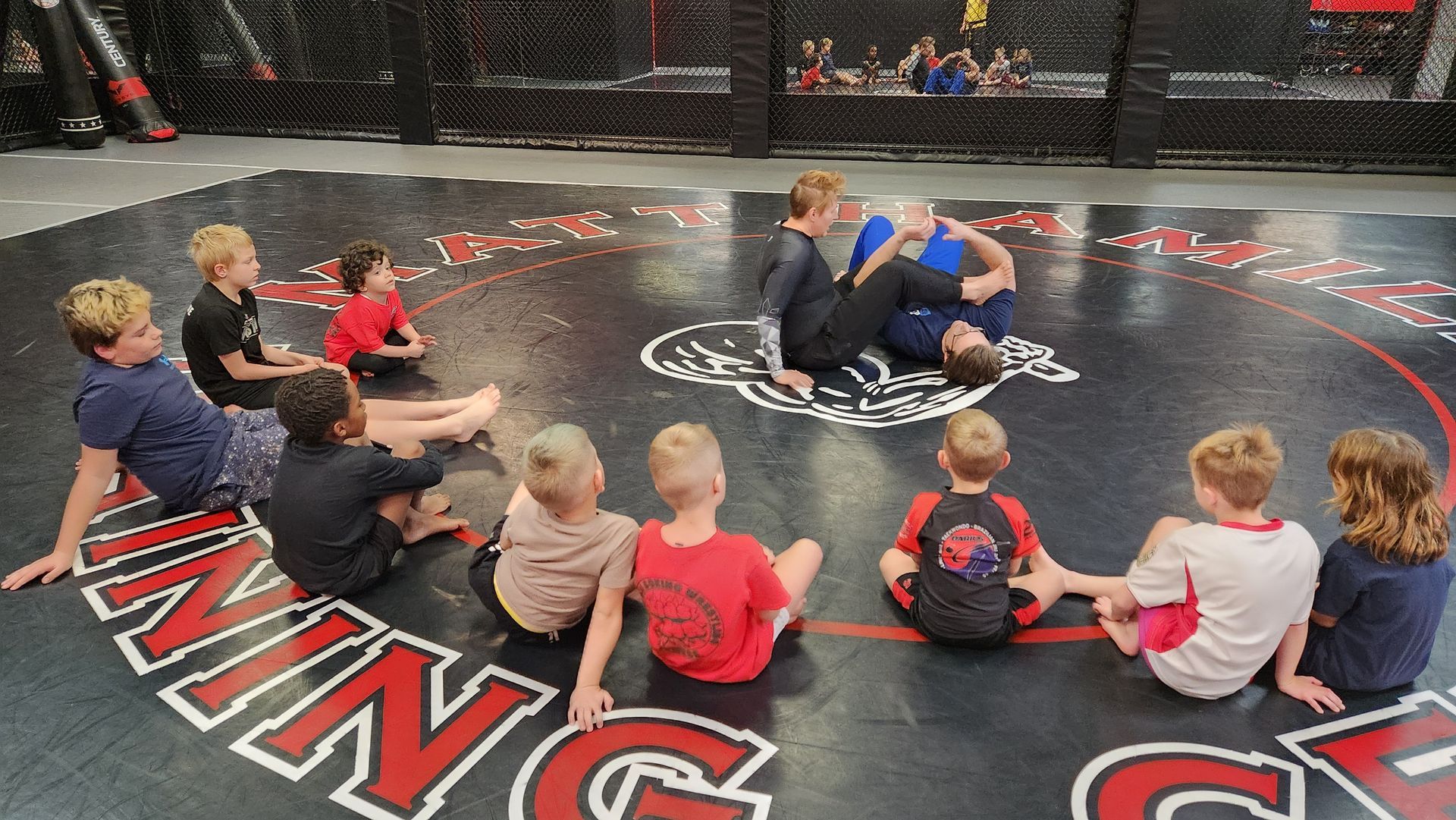 A group of children are sitting in a circle on a wrestling mat.