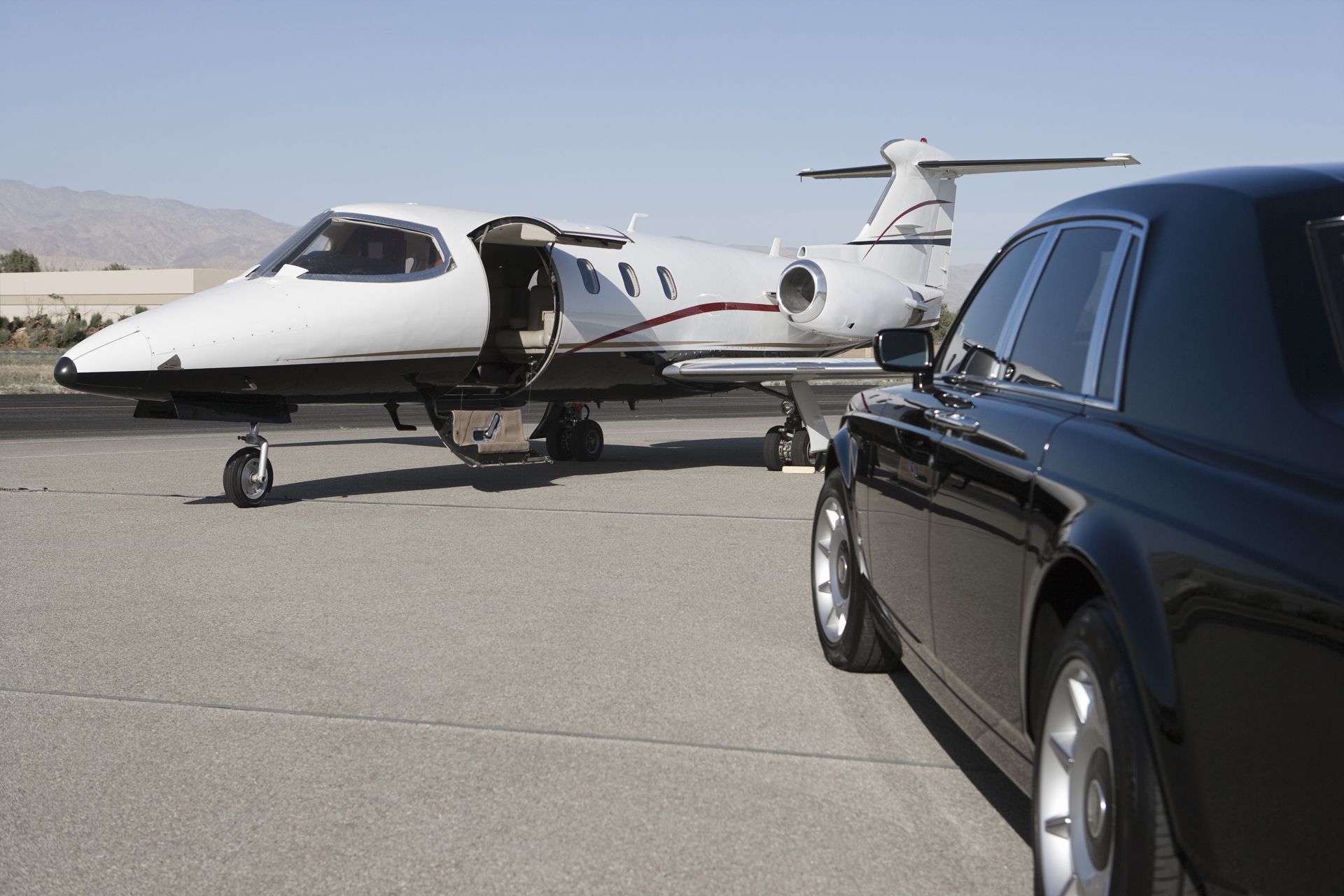 A private jet with its door open, next to a black luxury car on an airfield, bright sunlight
