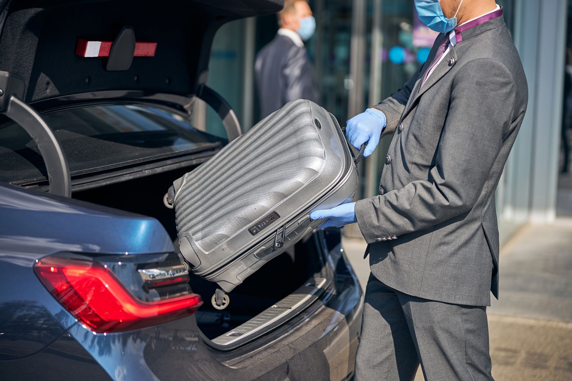 Man in suit with gloves loading suitcase into car trunk