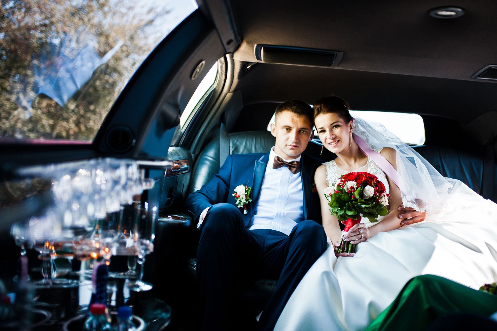 Bride and groom in formal wear seated inside a limousine, holding flowers, smiling