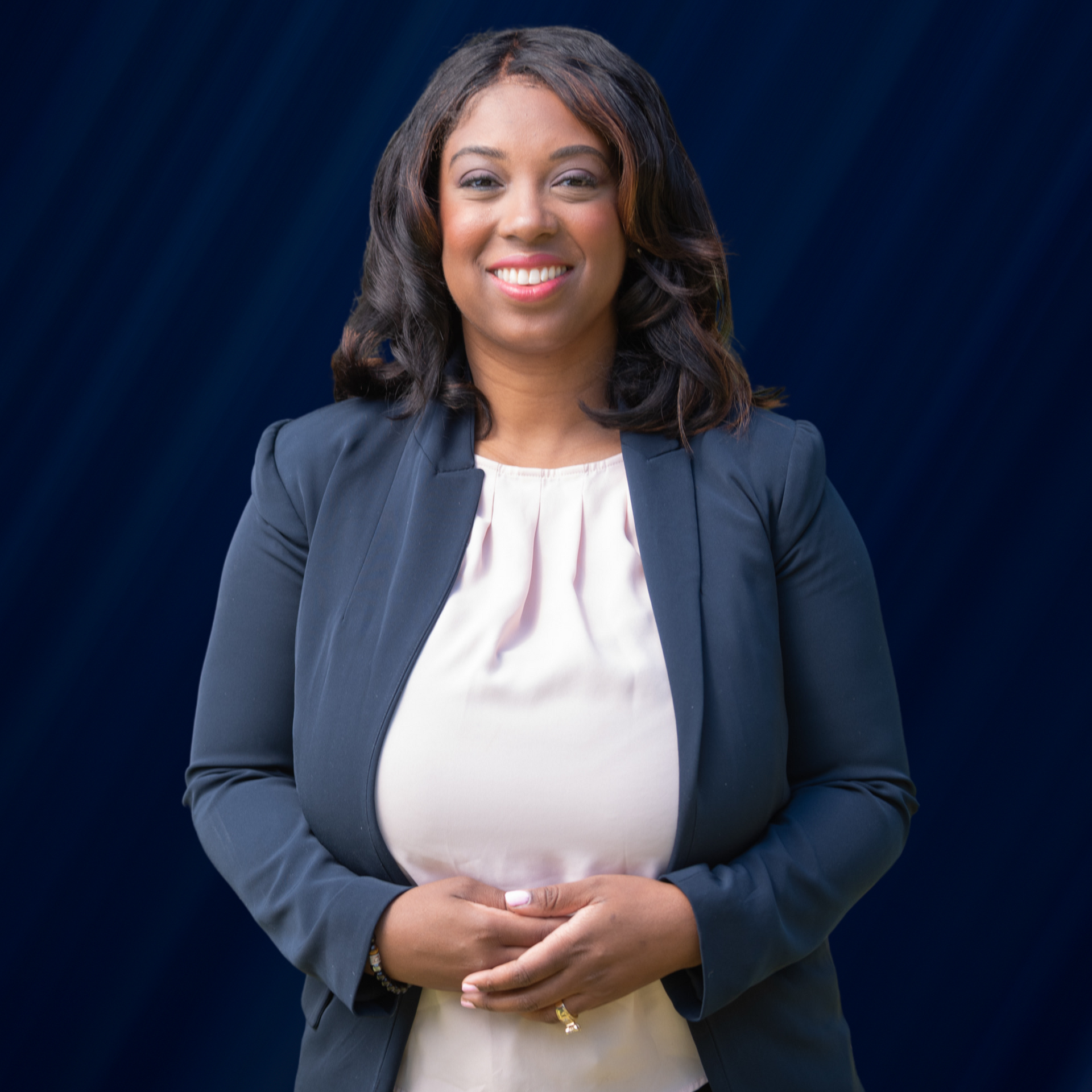 Woman in a navy blazer and pink blouse smiles, arms crossed, in front of a blue backdrop.