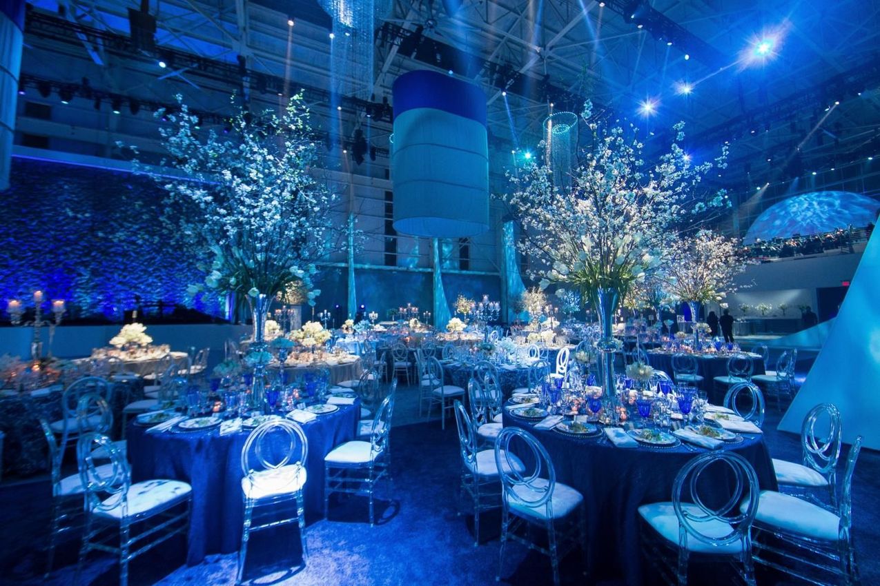Elegant event space with tables set for a formal dinner, illuminated by blue lighting and featuring floral centerpieces. 